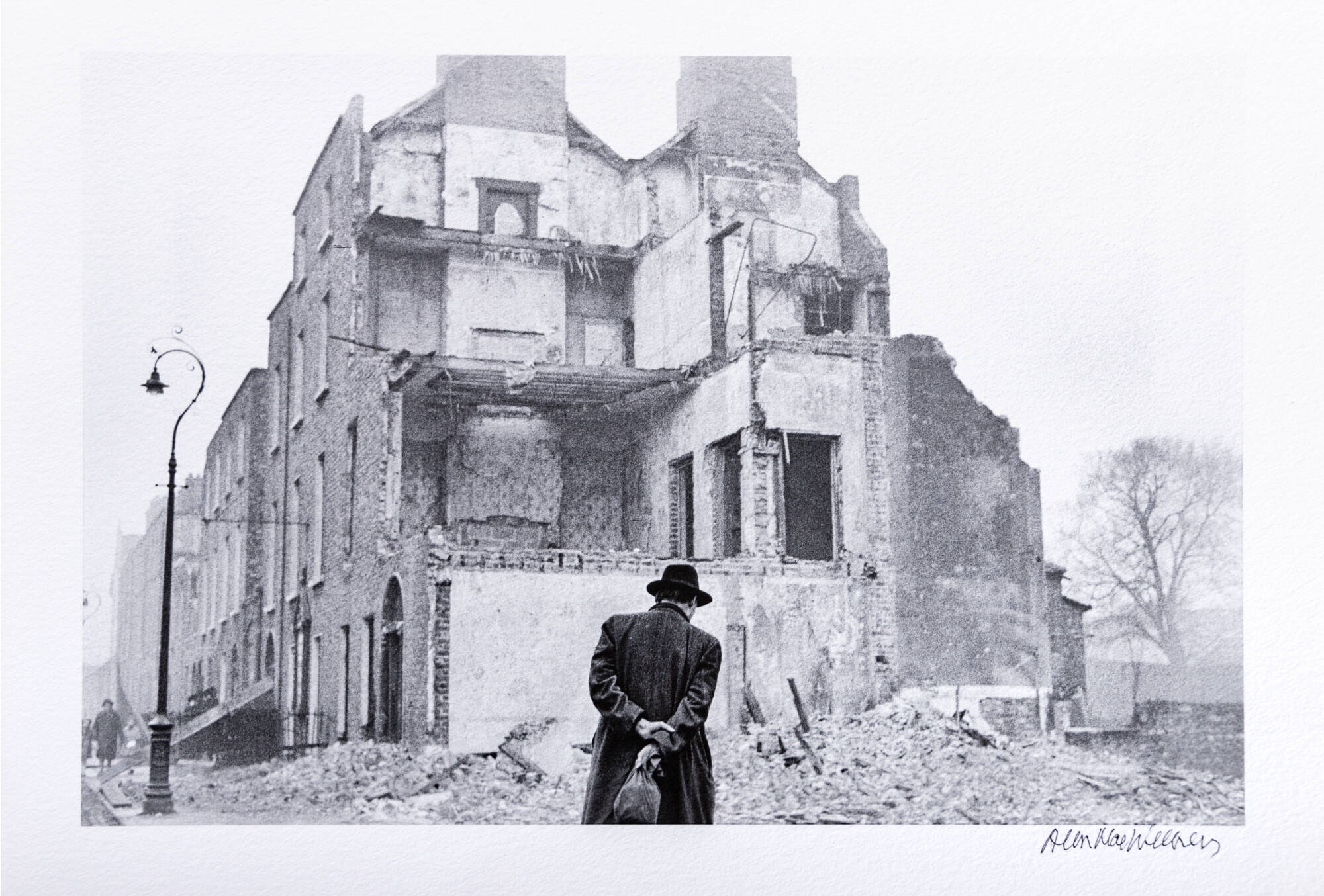 Black and white photograph of a man, standing with his hands behind his back, facing away from the viewer, looking at an old building.